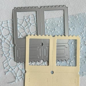 Op de afbeelding: Een zilverkleurige metalen snijmal in de vorm van een dubbel ticket met geschulpte randen en een centraal rond gat, geplaatst over een crèmekleurige kartonnen uitsnede van hetzelfde ontwerp, met zichtbare reliëfdetails en uitsparingen.
