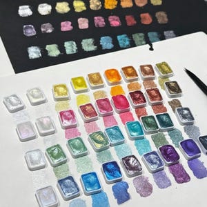 May include: A set of rectangular watercolour pans containing metallic and iridescent pigments in a wide spectrum of colours. Each pan is arranged on white paper with corresponding paint swatches beneath them, showing a shimmering finish.