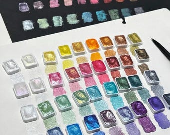 May include: A set of rectangular watercolor pans containing metallic and iridescent pigments in a wide spectrum of colors. Each pan is arranged on white paper with corresponding paint swatches beneath them, showing a shimmering finish.