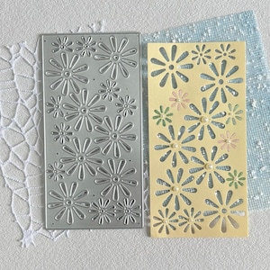Puede incluir: Un troquel rectangular de metal plateado con un patrón repetitivo de flores tipo margarita junto a un recorte de cartulina amarillo pálido. El recorte muestra el mismo diseño floral con pequeños adornos redondos de color blanco.