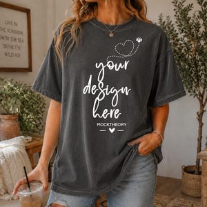 May include: A charcoal gray short-sleeve t-shirt featuring a white graphic print that reads "your design here" in a cursive script, accompanied by a dotted heart outline, a small bee icon, and the text "MOCKTHEORY" with a small heart symbol.