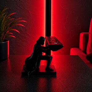 May include: A black Darth Vader figurine holding a TV remote. The figurine is on a black base, set against a red-lit background. A houseplant in a black pot is visible on the left.