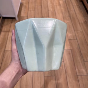 Könnte beinhalten: Ein blass mintgrüner Keramikübertopf mit einem geometrischen Design aus scharfen, vertikalen, kantigen Facetten. Der Topf hat eine breite, offene Oberseite und eine sich verjüngende Basis mit einer glatten, matten Oberfläche in hellem Grün.