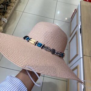 May include: A light beige straw sun hat with a wide brim, featuring a decorative band around the crown. The band consists of two rows of small turquoise, cream, and silver-toned beads, accented with a circular silver-toned metal charm.
