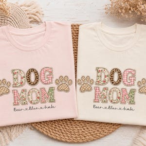 Könnte beinhalten: Ein hellrosa T-Shirt und ein cremefarbenes T-Shirt, beide mit dem Schriftzug DOG MOM in gemusterten Buchstaben und Pfotenabdrücken an den Seiten. Unter dem Text stehen die Namen Bear, Blue und Simba in Schreibschrift mit kleinen Herzsymbolen.