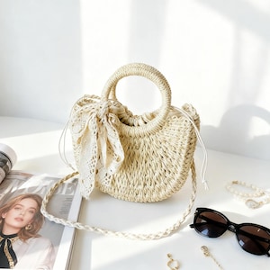 May include: A beige woven straw handbag with a semi-circular shape, featuring two round top handles, a drawstring closure, and a decorative cream-colored lace ribbon tied to one side. The bag includes a braided straw shoulder strap.