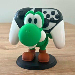 Puede incluir: Un soporte para mando con temática de Yoshi, verde, blanco y marrón. Sostiene un mando de videojuegos en blanco y negro. La figura de Yoshi tiene una cabeza verde, ojos blancos y zapatos marrones. El soporte está sobre una base circular negra.