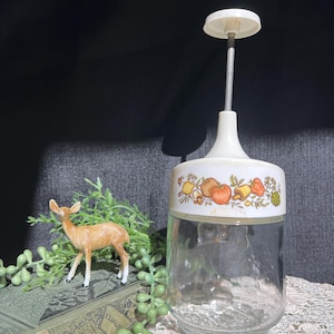 May include: A vintage white and glass food chopper with a decorative band featuring colorful illustrations of vegetables. The chopper has a clear glass jar base and a white handle. A small deer figurine and greenery are in the background.