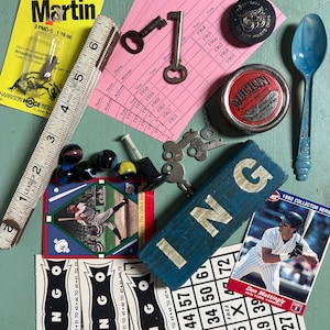 May include: A collection of vintage items including a Panther Martin fishing lure package, a wooden ruler, pink ledger cards, metal keys, a round Mucilin tin, a blue plastic spoon, marbles, a blue block with letters, and two baseball cards.