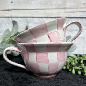 May include: Two ceramic cups stacked on top of each other, each featuring a hand-painted light pink and white checkered pattern. The cups have a wide, rounded bowl shape with a curved handle on the side and a smooth, glossy finish throughout.