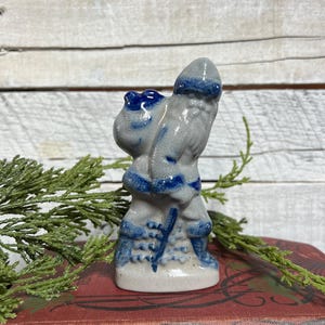 May include: A small ceramic figurine of a bearded figure wearing a hat and boots, carrying a large sack on their back and holding a staff. The piece features a light gray base glaze with dark blue painted accents on the hat, sack, and boots.