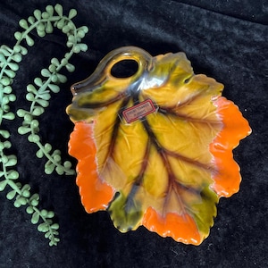 May include: A ceramic leaf-shaped dish with a handle, featuring a vibrant color palette of yellow, orange, and green. The dish has a glossy finish and a detailed vein pattern. A small label is visible. Decorative item.