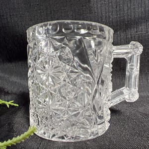 May include: Clear glass mug with a handle. The mug features a detailed, geometric pattern across its surface. The handle is also clear and has a decorative design. The mug is sitting on a dark surface.