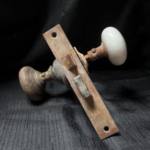 May include: Antique door hardware set featuring a rusty metal plate and two door knobs. One knob is a white, cracked porcelain oval, the other is a round, aged metal knob. The set includes a latch mechanism.