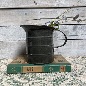 May include: A vintage green enamel pitcher with a handle, sitting on top of a book titled "Fifty Fairy Tales" by Whitman. The pitcher is filled with dried blue flowers.