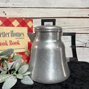 May include: A vintage aluminum percolator coffee pot with a black plastic handle and a matching black knob on the lid. The pot has a wide base and a tapered body. Next to it is a Better Homes and Gardens Cook Book with a red and yellow checkered cover.