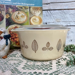 May include: A cream-colored Pyrex bowl with a clear lid, featuring a brown leaf pattern. The bowl is on a lace tablecloth, with a cookbook and a ceramic goose figurine in the background. The bowl is designed for food storage.