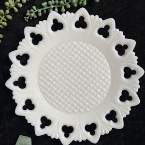 May include: A round white milk glass plate featuring a central diamond-patterned texture and a decorative openwork border with repeating clover-shaped cutouts. The plate has a scalloped edge and a smooth, glossy finish throughout the surface.