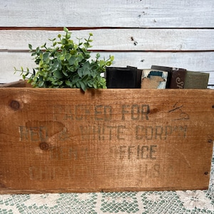 May include: A rustic wooden crate with printed text, filled with small books and green foliage. The crate has a weathered appearance, with visible wood grain and knots. The text on the crate reads "PACKED FOR... WHITE CORP... OFFICE... U.S."