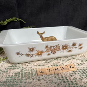 May include: White Pyrex baking dish with a brown floral design and a small deer figurine inside. The dish has a rectangular shape with handles and is set on a lace tablecloth.