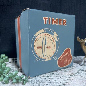 May include: A vintage Mirro-Matic kitchen timer in its original box. The box is light blue with orange accents and features the word "TIMER" in red. The timer's face is illustrated on the box, along with the brand name. A small deer figurine is in the background.