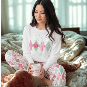 Op de afbeelding: Een wit shirt met lange mouwen voorzien van een centrale rij ruitvormen in roze, groen en wit met bloemen- en ruitpatronen, gecombineerd met een bijpassende pyjamabroek met een herhalend ruitpatroon in roze, groen en witte kleuren.