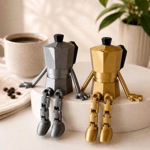May include: Two articulated figures shaped like moka pot coffee makers, one in metallic silver and one in metallic gold. Each figure features a faceted body, a black knob on the lid, and jointed arms and legs that allow them to sit upright.