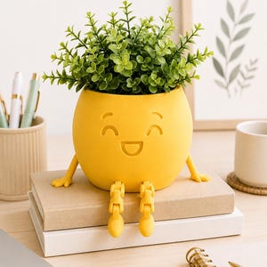 May include: A bright yellow planter shaped like a round character with a smiling face, small arms, and dangling articulated legs. The planter holds a lush green artificial plant with small leaves, creating a cheerful and functional desk accessory.