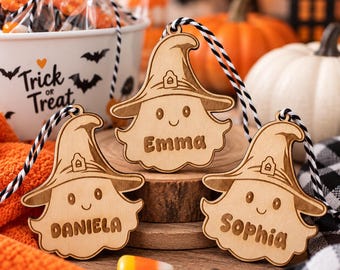 May include: Three light-colored wooden ornaments shaped like ghosts wearing witch hats, each engraved with the names Emma, Daniela, or Sophia. Each ornament features a black and white striped string loop for hanging and a smiling face.