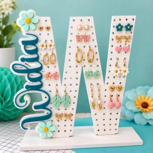 May include: A white letter W earring holder with a teal script name plate reading "Widia" and a teal flower accent. The stand features numerous holes for organizing various stud and dangle earrings, including heart, butterfly, bow, and leaf shapes.