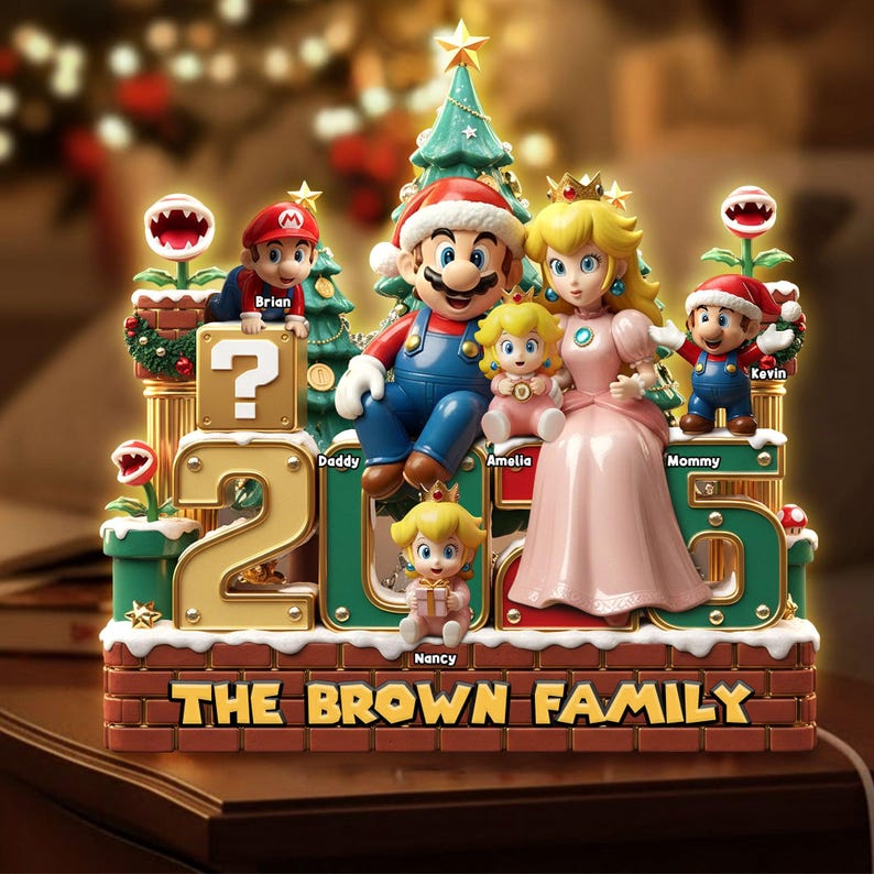 Może przedstawiać: Ozdobna figurka rodzinna z motywem Super Mario przedstawiająca Mario, księżniczkę Peach i mniejsze postacie. Podstawa zawiera napis THE BROWN FAMILY oraz imiona Brian, Daddy, Amelia, Mommy i Nancy przy postaciach i rok 2025.