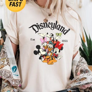 K&ouml;nnte beinhalten: Ein cremefarbenes Kurzarm-T-Shirt mit einem Grafikdruck von Mickey Mouse, Minnie Mouse, Donald Duck, Daisy Duck, Goofy und Pluto. Der Text lautet Disneyland Est 1955. Das T-Shirt hat einen Rundhalsausschnitt und eine lockere Passform.