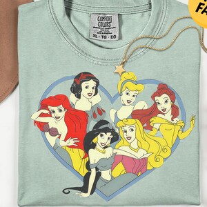 May include: A sage green crew neck t-shirt featuring a graphic print of six animated princesses arranged inside a blue heart outline. The shirt has a Comfort Colors brand label at the inner neckline indicating size XL and is made of 100% cotton.