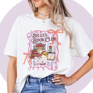 Può includere: Una t-shirt bianca a maniche corte con una stampa grafica che riporta il testo Belle's Book Club, Est. 1991 e Tale As Old As Time. Il design include una pila di libri, una rosa sotto una cupola di vetro e un personaggio teiera con tazza.