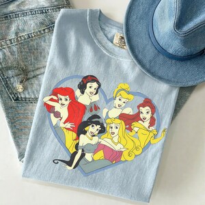 May include: A light blue t-shirt featuring a graphic print of six animated princesses arranged inside a heart shape. The characters include Ariel, Snow White, Cinderella, Jasmine, Aurora, and Belle. The shirt has a crew neckline and a tag.