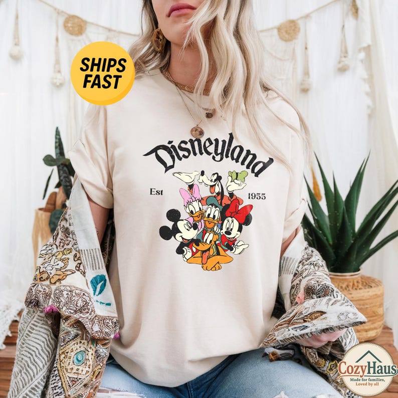 K&ouml;nnte beinhalten: Ein cremefarbenes Kurzarm-T-Shirt mit einem Grafikdruck von Mickey Mouse, Minnie Mouse, Donald Duck, Daisy Duck, Goofy und Pluto. Der Text lautet Disneyland Est 1955. Das T-Shirt hat einen Rundhalsausschnitt und eine lockere Passform.