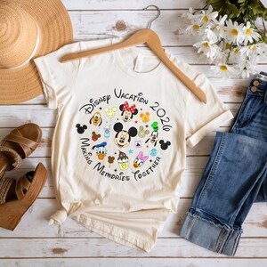 May include: A cream-colored short-sleeve t-shirt featuring a circular graphic with Disney characters, icons, and the text Disney Vacation 2020 Making Memories Together. The shirt is displayed with a straw hat, blue jeans, and brown wedge sandals.