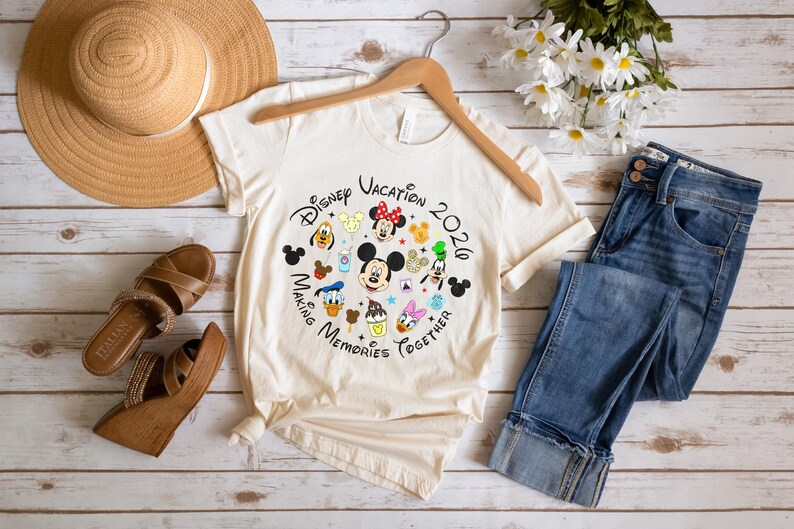 May include: A cream-colored short-sleeve t-shirt featuring a circular graphic with Disney characters, icons, and the text Disney Vacation 2020 Making Memories Together. The shirt is displayed with a straw hat, blue jeans, and brown wedge sandals.