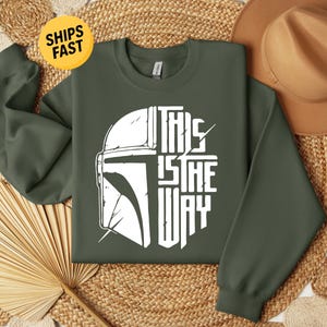 May include: An olive green crewneck sweatshirt featuring a white graphic print on the chest. The design displays the left half of a helmet and the text "THIS IS THE WAY" on the right half, arranged in a circular shape with distressed details.