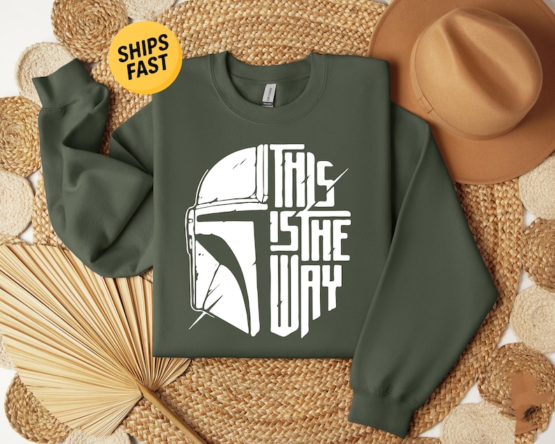 May include: An olive green crewneck sweatshirt featuring a white graphic print on the chest. The design displays the left half of a helmet and the text "THIS IS THE WAY" on the right half, arranged in a circular shape with distressed details.
