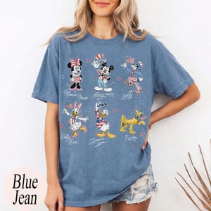 May include: A blue short-sleeve t-shirt featuring a graphic print of Disney characters including Minnie Mouse, Mickey Mouse, Goofy, Daisy Duck, Donald Duck, and Pluto, all dressed in patriotic red, white, and blue outfits with small stars.