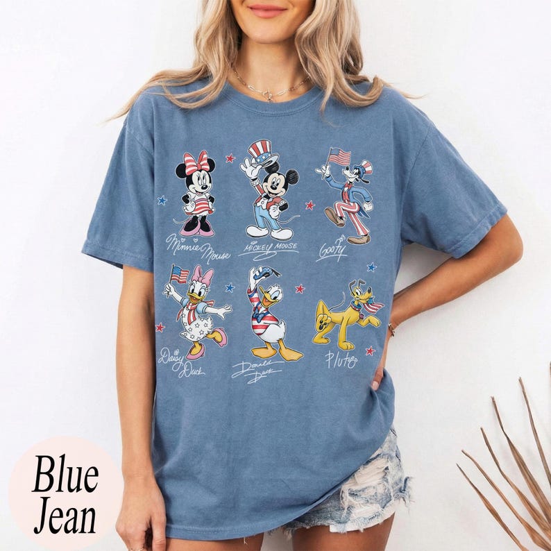 May include: A blue short-sleeve t-shirt featuring a graphic print of Disney characters including Minnie Mouse, Mickey Mouse, Goofy, Daisy Duck, Donald Duck, and Pluto, all dressed in patriotic red, white, and blue outfits with small stars.