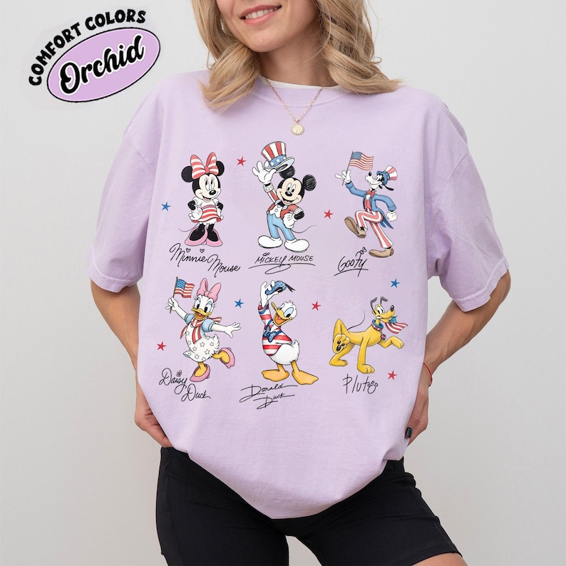 May include: A light purple t-shirt featuring a graphic print of six cartoon characters: Minnie Mouse, Mickey Mouse, Goofy, Daisy Duck, Donald Duck, and Pluto. Each character is dressed in patriotic red, white, and blue clothing with stars and stripes.