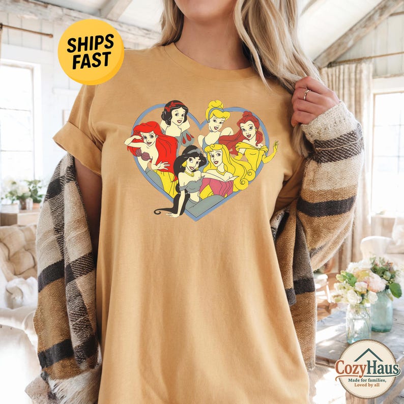 May include: A mustard yellow short-sleeve t-shirt featuring a graphic print of six animated princesses arranged inside a blue heart outline. The characters include figures with red, black, blonde, and brown hair wearing colorful dresses.
