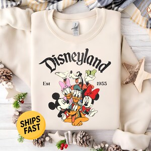 K&ouml;nnte beinhalten: Ein cremefarbenes Sweatshirt mit Rundhalsausschnitt und einem schwarzen Aufdruck des Disneyland-Schriftzugs, Est 1955 sowie einer bunten Illustration von Mickey Maus, Minnie Maus, Donald Duck, Daisy Duck, Goofy und Pluto in einer Gruppe.