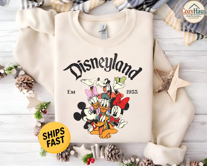 K&ouml;nnte beinhalten: Ein cremefarbenes Sweatshirt mit Rundhalsausschnitt und einem schwarzen Aufdruck des Disneyland-Schriftzugs, Est 1955 sowie einer bunten Illustration von Mickey Maus, Minnie Maus, Donald Duck, Daisy Duck, Goofy und Pluto in einer Gruppe.