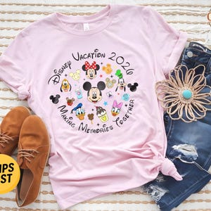 May include: A light pink t-shirt featuring a circular graphic with the text Disney Vacation 2026 Making Memories Together, surrounded by illustrations of Mickey Mouse, Minnie Mouse, Donald Duck, Daisy Duck, Goofy, Pluto, and various snacks.