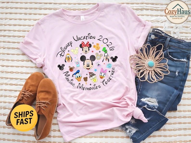 May include: A light pink t-shirt featuring a circular graphic with the text Disney Vacation 2026 Making Memories Together, surrounded by illustrations of Mickey Mouse, Minnie Mouse, Donald Duck, Daisy Duck, Goofy, Pluto, and various snacks.