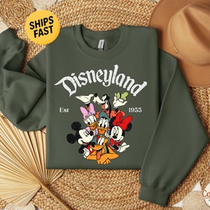 K&ouml;nnte beinhalten: Ein olivgr&uuml;nes Sweatshirt mit Rundhalsausschnitt und einem wei&szlig;en Aufdruck des Disneyland-Schriftzugs, Est 1955 sowie einer bunten Illustration von Mickey Maus, Minnie Maus, Donald Duck, Daisy Duck, Goofy und Pluto in einer Gruppe.