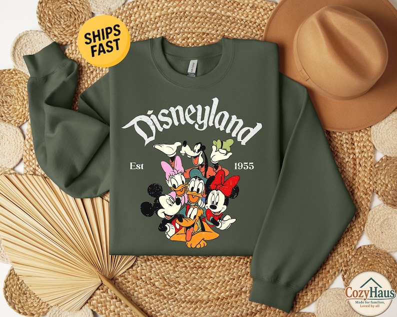 K&ouml;nnte beinhalten: Ein olivgr&uuml;nes Sweatshirt mit Rundhalsausschnitt und einem wei&szlig;en Aufdruck des Disneyland-Schriftzugs, Est 1955 sowie einer bunten Illustration von Mickey Maus, Minnie Maus, Donald Duck, Daisy Duck, Goofy und Pluto in einer Gruppe.